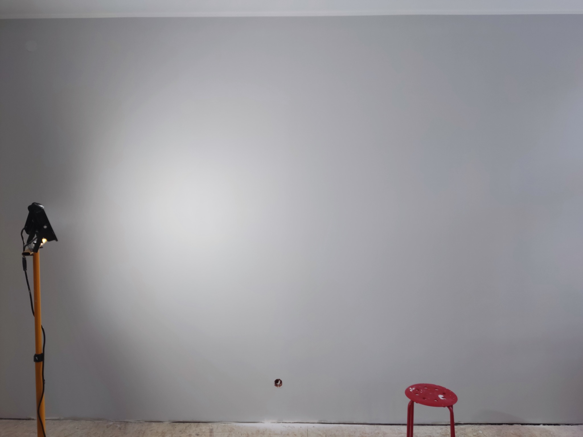 Świeżo pomalowana szara ściana oświetlona lampą halogenową na drewnianym statywie, mały czerwony stołek w rogu, widoczne gniazdko elektryczne.