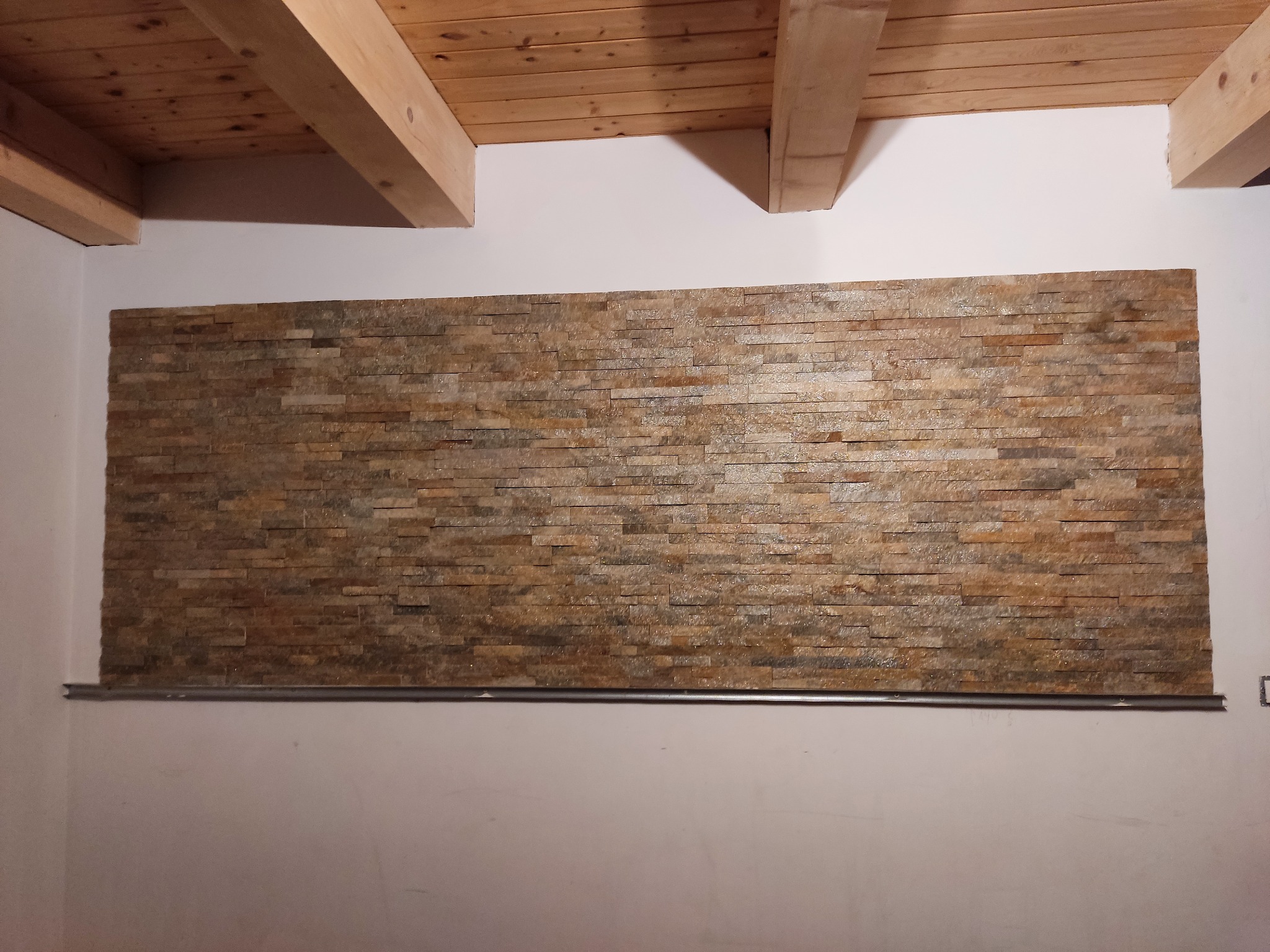Fragment ściany z dekoracyjną okładziną kamienną w odcieniach brązu i beżu, zamontowaną na metalowym profilu, widoczny drewniany sufit z belkami.