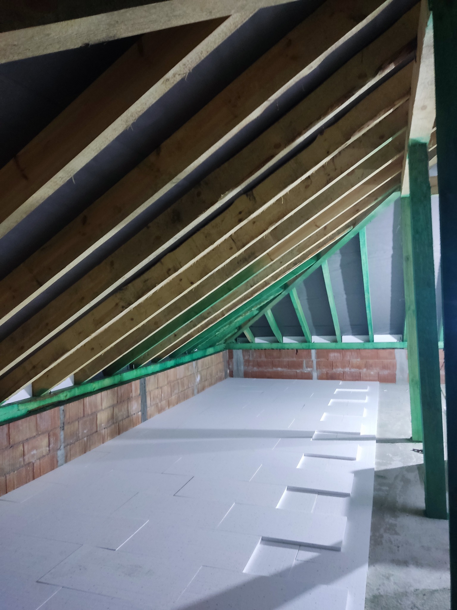 Ocieplenie poddasza: białe płyty styropianowe ułożone na podłodze, widoczne drewniane krokwie dachu i zielona konstrukcja wzmacniająca.