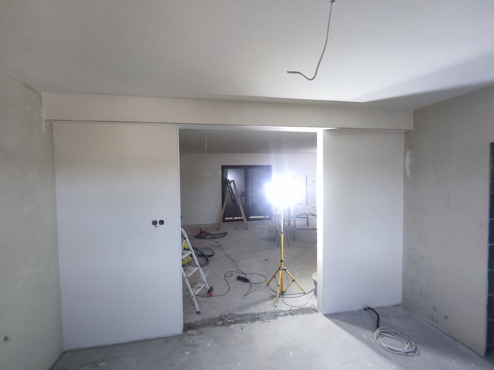 Wnętrze w trakcie remontu: widoczne gładzie na ścianach, drabina, lampa budowlana na statywie oraz przewody elektryczne wystające z sufitu.