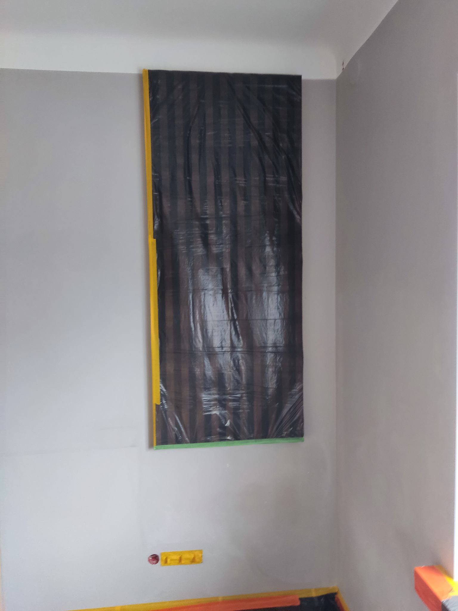 Przygotowanie do prac malarskich: ściana zabezpieczona folią malarską z żółtą taśmą, widoczne gniazdko elektryczne również zabezpieczone taśmą.