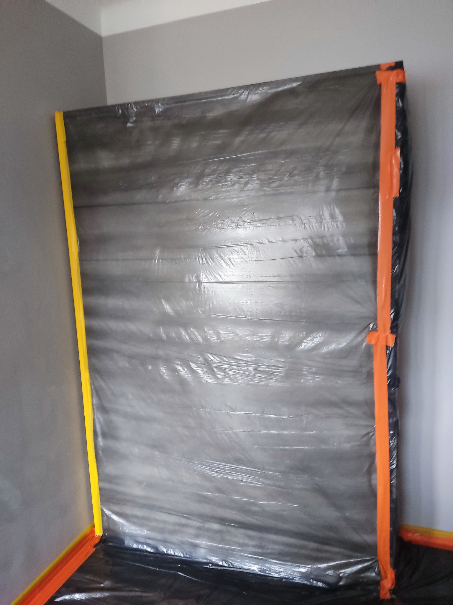 Ściana zabezpieczona folią ochronną i taśmą malarską w kolorach żółtym i pomarańczowym przed pracami wykończeniowymi, widoczne szare ściany w tle.