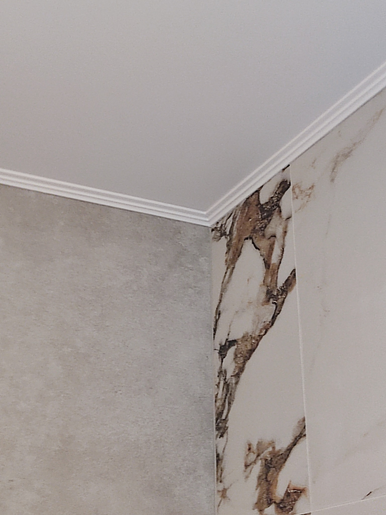 Fragment wykończonego narożnika: gładka, szara ściana sąsiaduje z płytkami imitującymi marmur, a sufit zdobi biała listwa dekoracyjna.