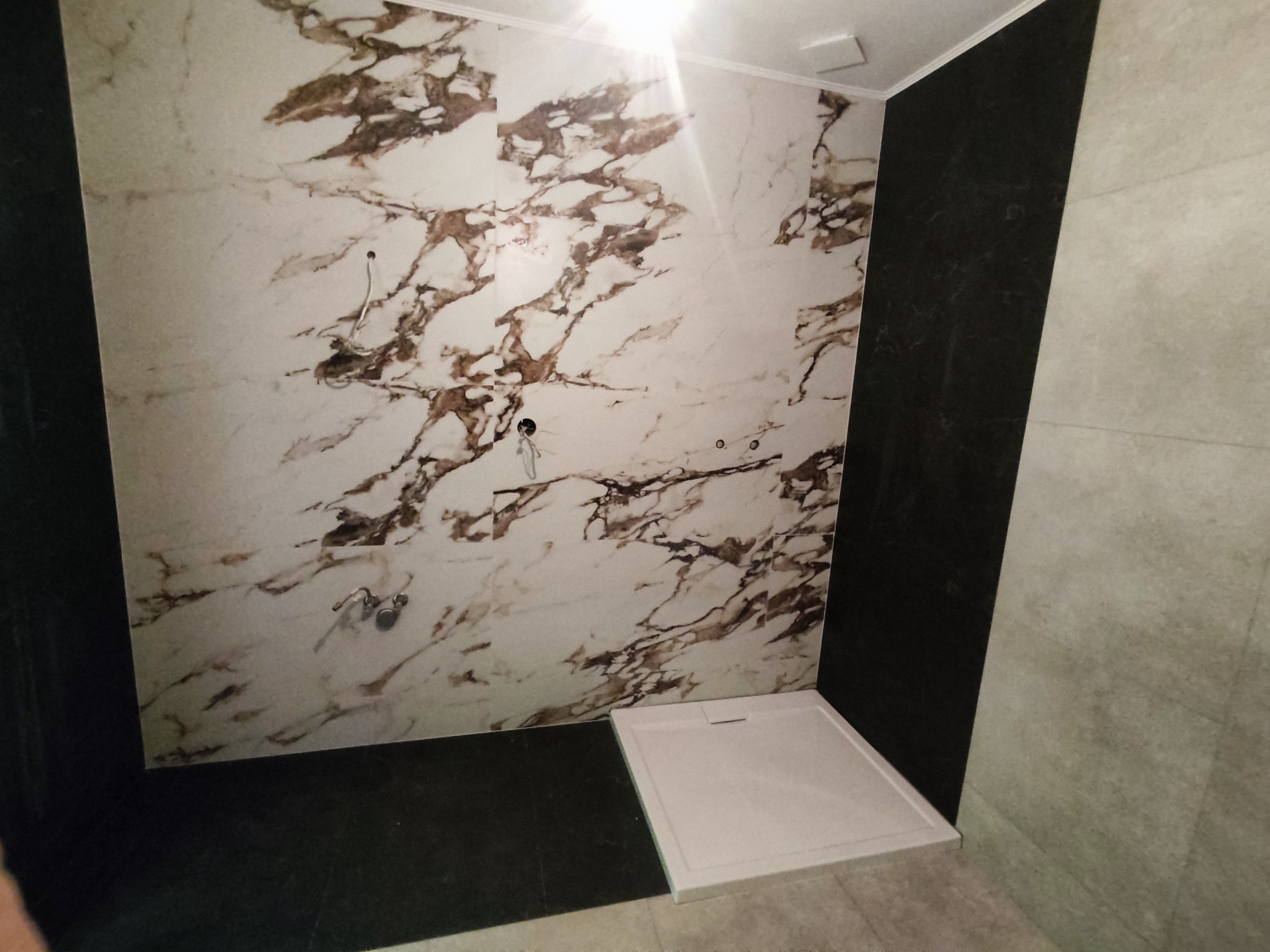 Nowo wykończona kabina prysznicowa z panelami ściennymi imitującymi marmur i czarnym panelem obok, widoczne wystające kable i rury przed montażem armatury.
