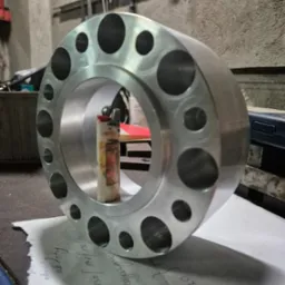 Aluminiowy pierścień z otworami na tle warsztatu, z zapalniczką wewnątrz, leżący na kartce z odręcznymi notatkami.