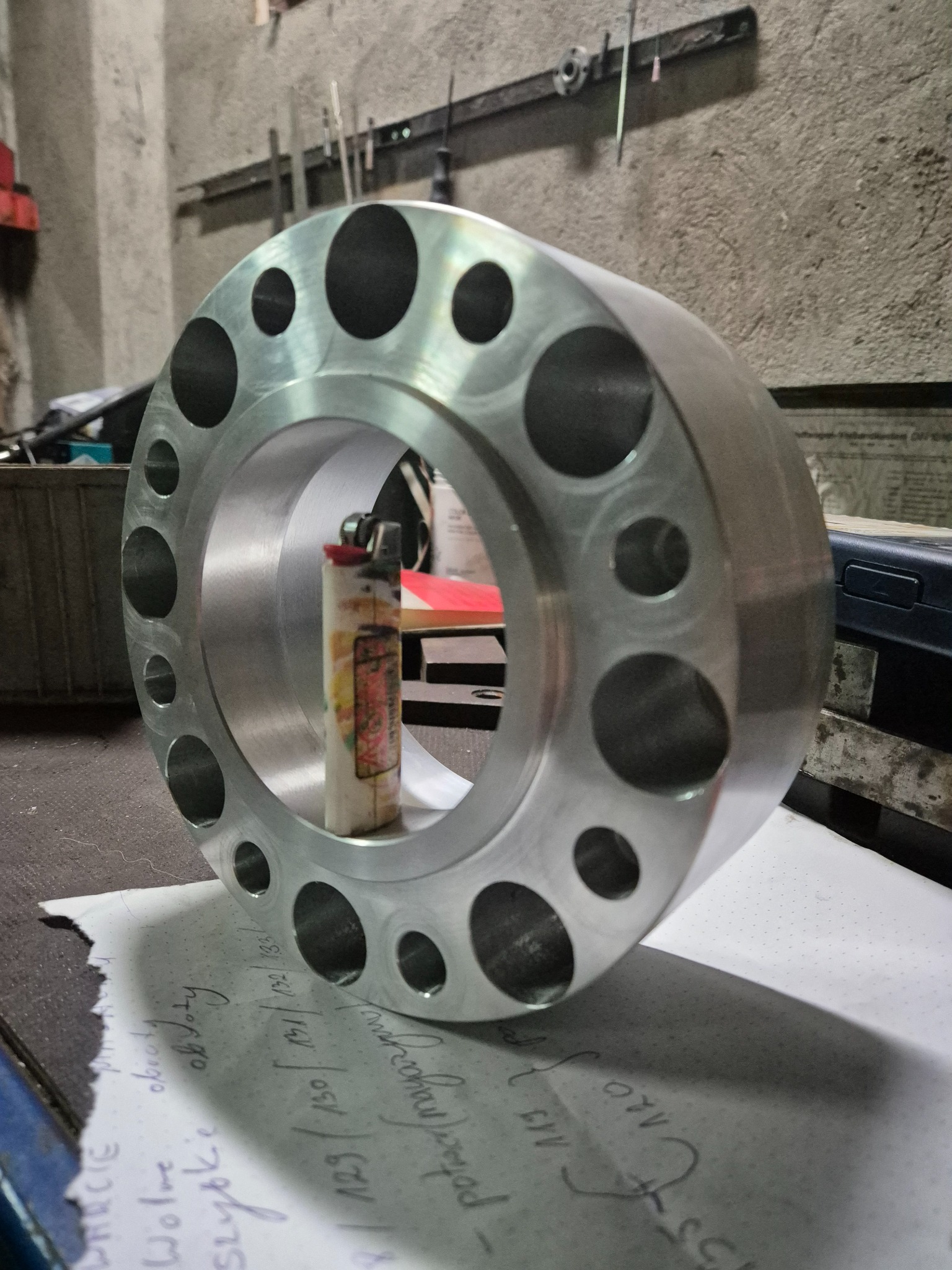 Aluminiowy pierścień z otworami na tle warsztatu, z zapalniczką wewnątrz, leżący na kartce z odręcznymi notatkami.