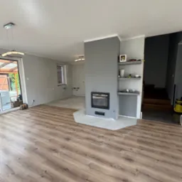 Salon po remoncie: drewniana podłoga, szary kominek z półkami, częściowo wykończona ściana w tle. Widoczne schody i wyjście na zewnątrz.