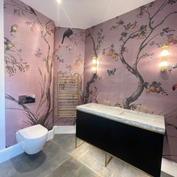 Łazienka i Dom Melchior Nowociński - Stylowa łazienka z tapetą w motywy drzew i ptaków. Czarna szafka z marmurowym blatem, złoty grzejnik i szara podłoga tworzą elegancki wystrój.