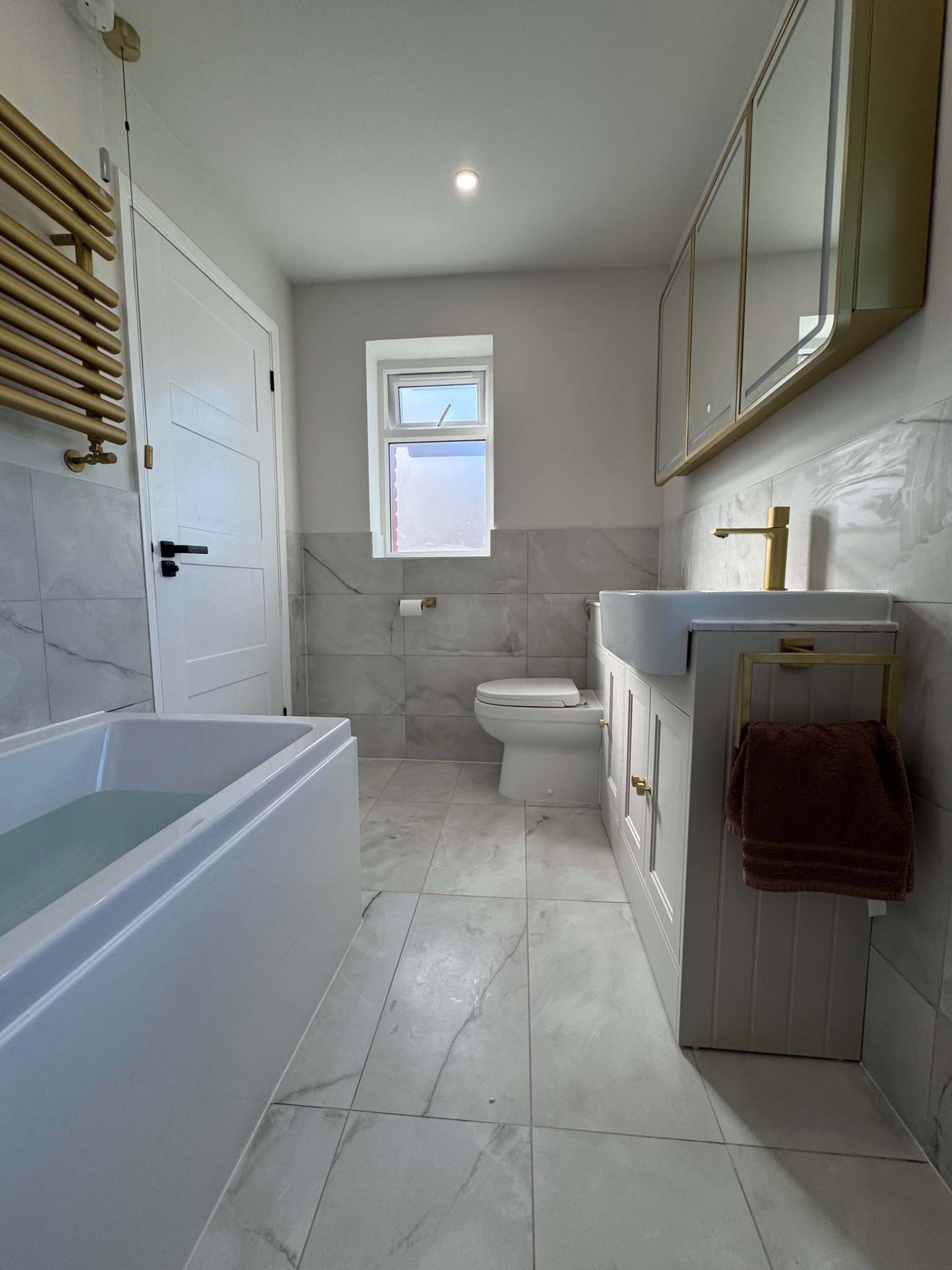 Jasna łazienka po remoncie: marmurowe płytki, biała wanna z wodą, umywalka z szafką, złota armatura i grzejnik drabinkowy. Nowoczesne wykończenie.