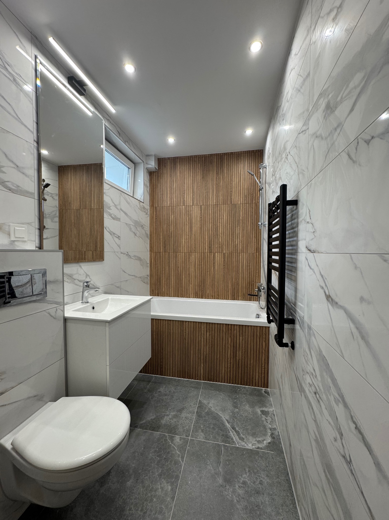 Nowoczesna łazienka: białe płytki imitujące marmur, drewniane panele przy wannie, szara podłoga, biała umywalka i podświetlane lustro.
