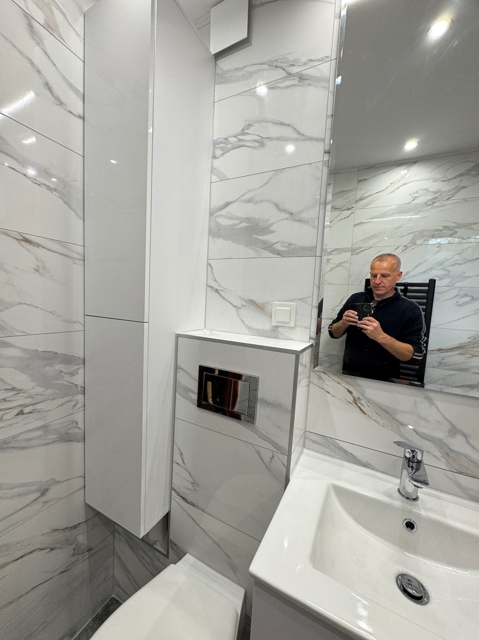 Biała łazienka z marmurowymi płytkami, wiszącą szafką i toaletą. W lustrze odbija się osoba robiąca zdjęcie. Widoczna umywalka i bateria.