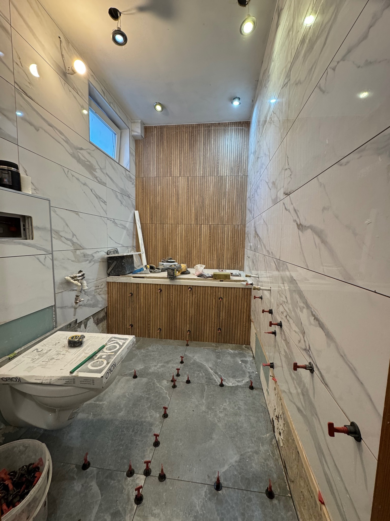 Łazienka w trakcie wykańczania w Miechucinie: marmurowe płytki na ścianach, drewnopodobne panele, poziomowanie płytek na podłodze, narzędzia glazurnicze.