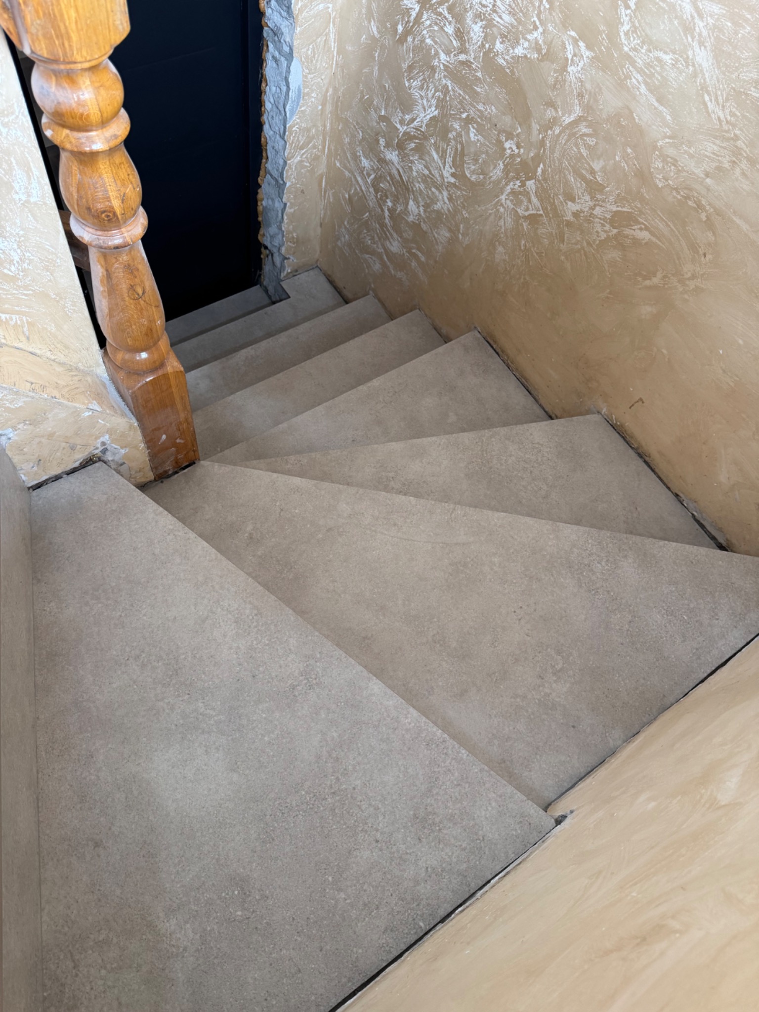 Wykończone schody z szarymi płytkami, z drewnianą balustradą i beżową ścianą z dekoracyjną strukturą. Widok z góry na fragment klatki schodowej po remoncie.