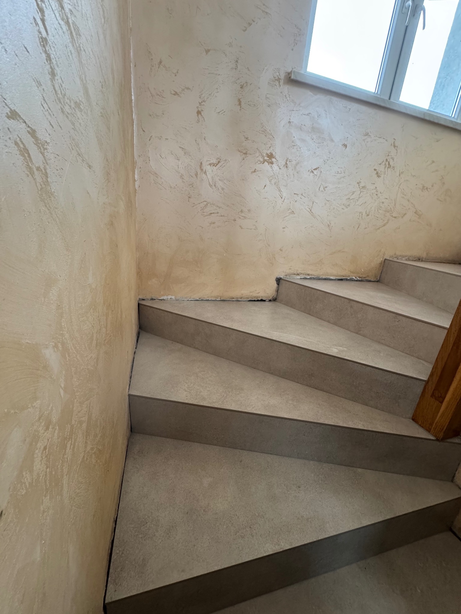 Wykończone schody z szarymi płytkami, widoczne dekoracyjne wykończenie ścian w odcieniach beżu i okno w tle. Ukończony etap prac.
