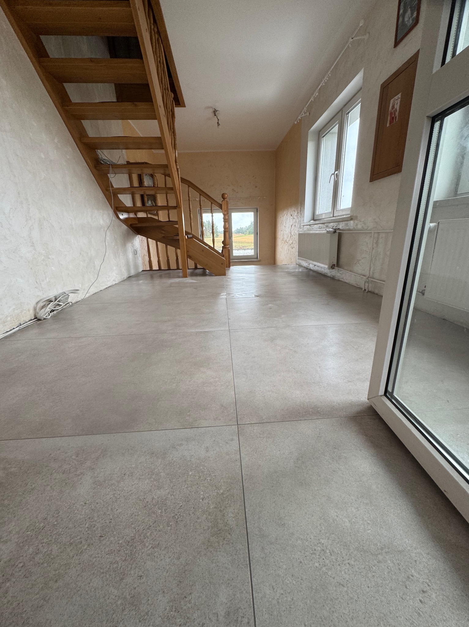 Wnętrze po remoncie: jasnoszara podłoga z płytek, drewniane schody, okna i grzejnik. Widok na zewnątrz przez okno. Minimalistyczny styl.