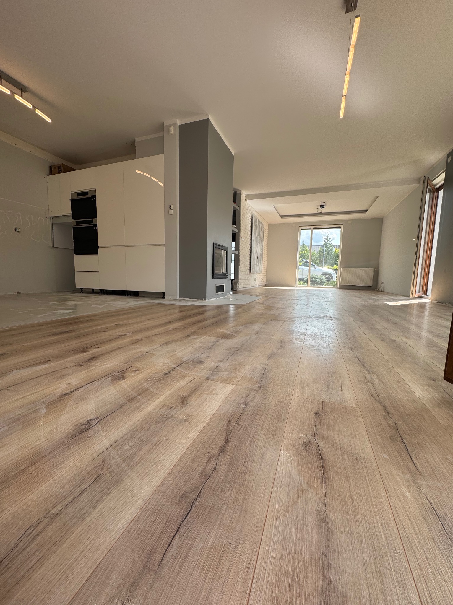 Jasne panele podłogowe w przestronnym, nowoczesnym wnętrzu z białymi meblami kuchennymi i szarą ścianą z kominkiem. Duże okno wpuszcza naturalne światło.