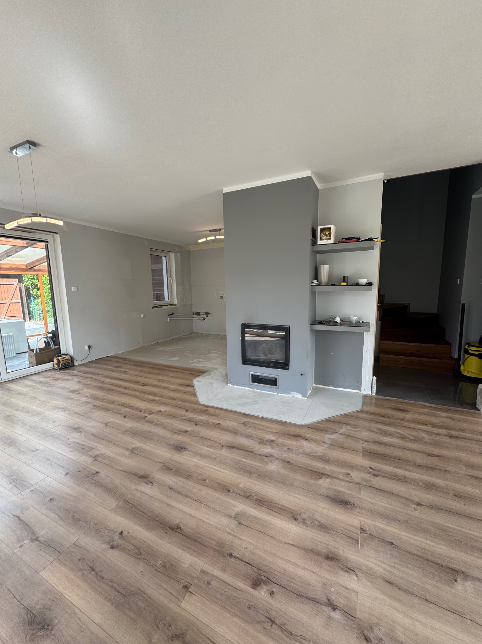 Salon po remoncie: drewniana podłoga, szary kominek z półkami, częściowo wykończona ściana w tle. Widoczne schody i wyjście na zewnątrz.