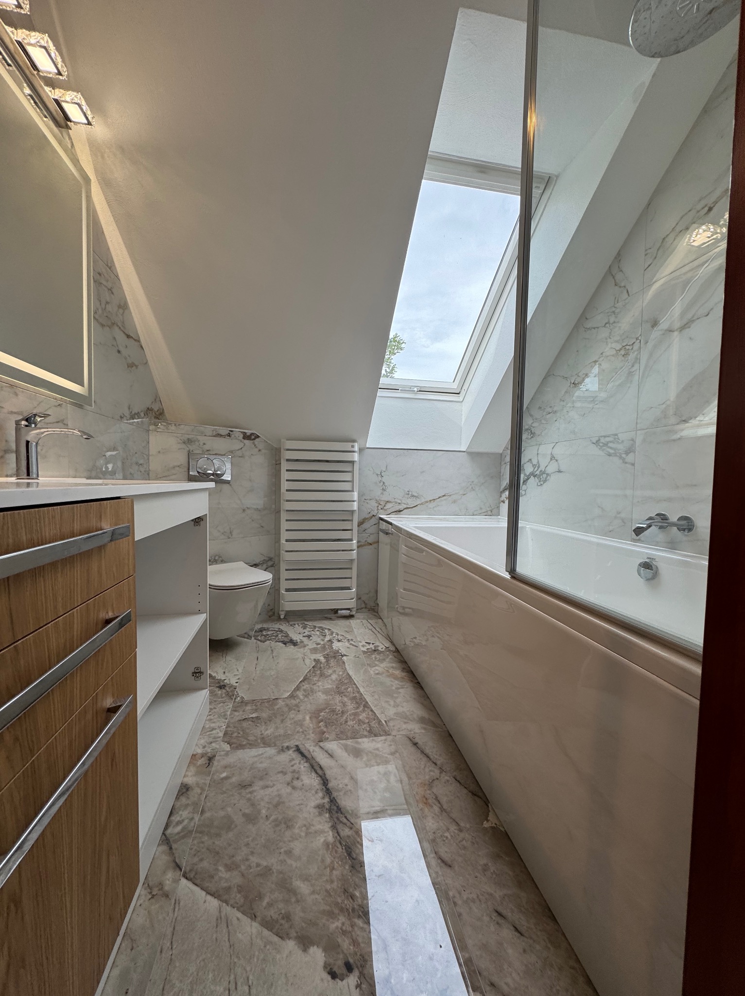 Wykończona łazienka na poddaszu z marmurowymi płytkami, oknem dachowym i grzejnikiem drabinkowym. Umywalka z szafką, toaleta i wanna za szklaną ścianką.