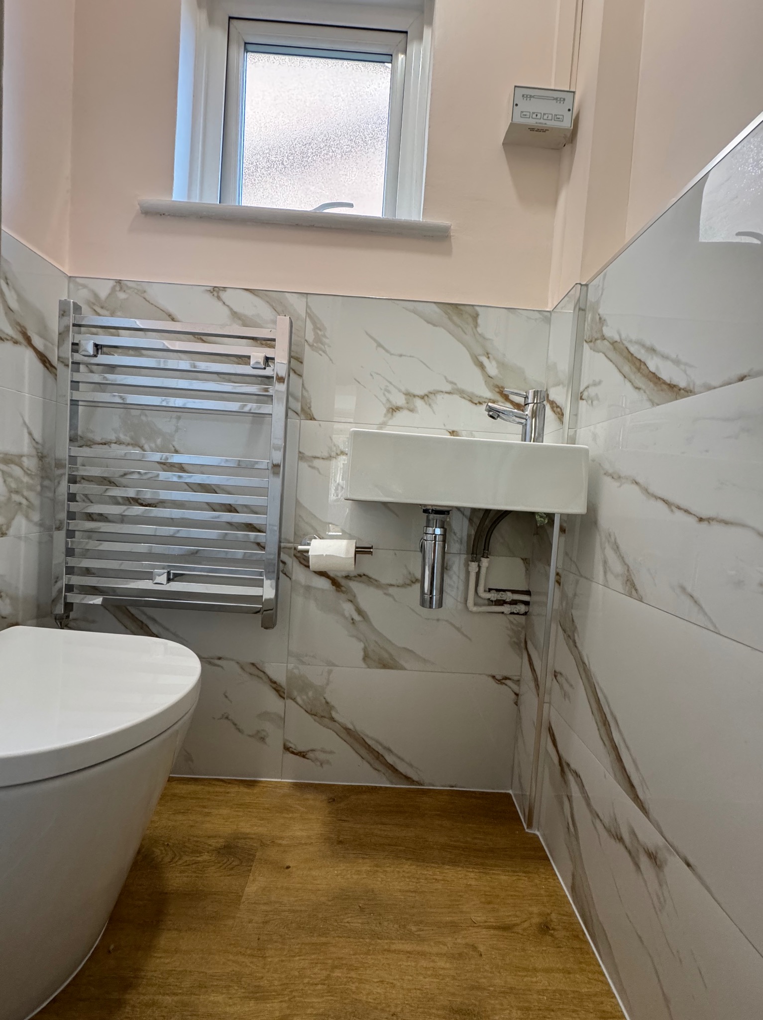 Wykończona łazienka z marmurowymi płytkami, wiszącą umywalką, grzejnikiem drabinkowym i drewnopodobną podłogą. Widok na fragment toalety.
