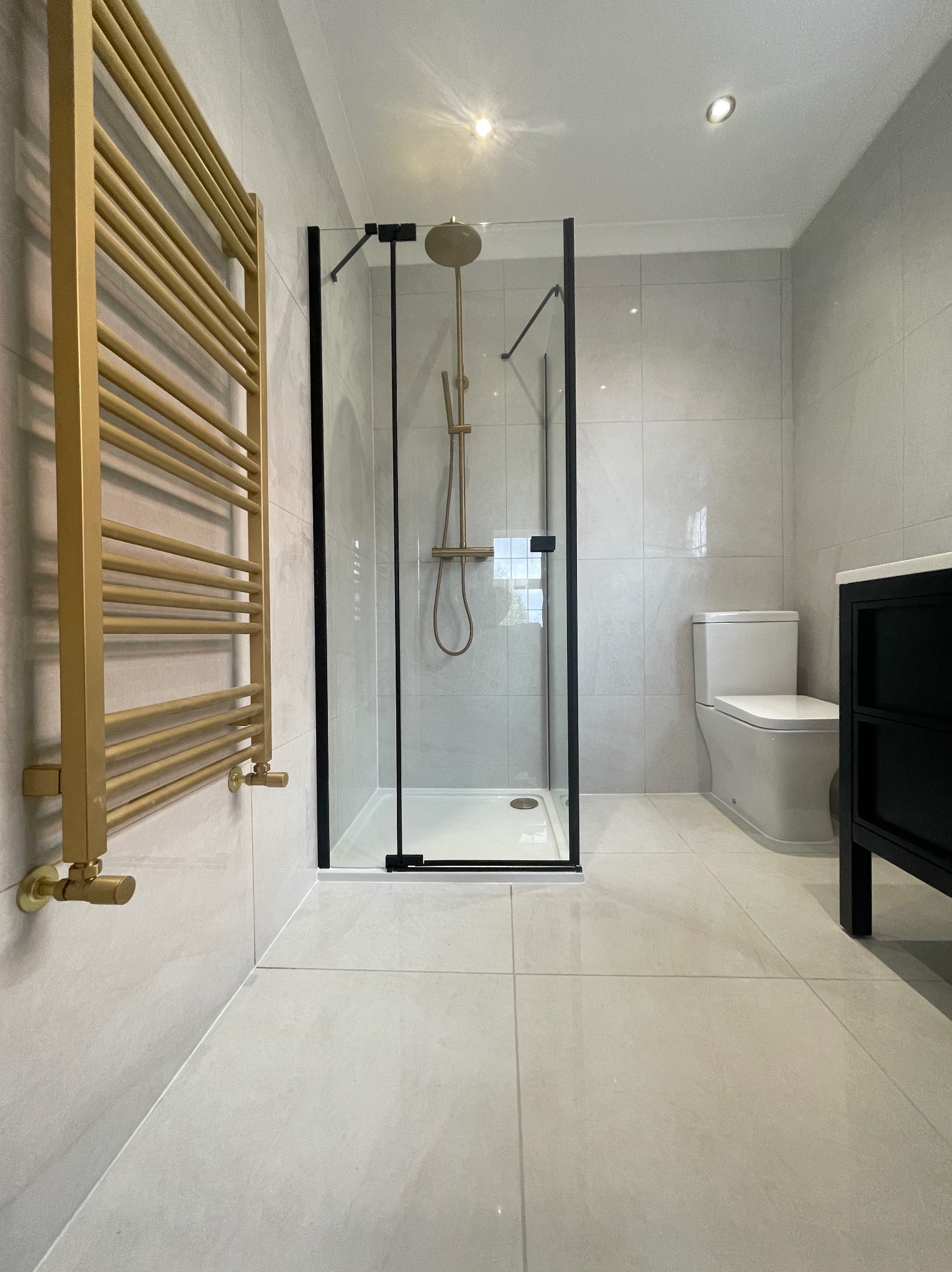 Nowoczesna łazienka z kabiną prysznicową w czarnej ramie, złotym grzejnikiem drabinkowym i dużymi, jasnymi płytkami. Minimalistyczny design i stonowana kolorystyka.