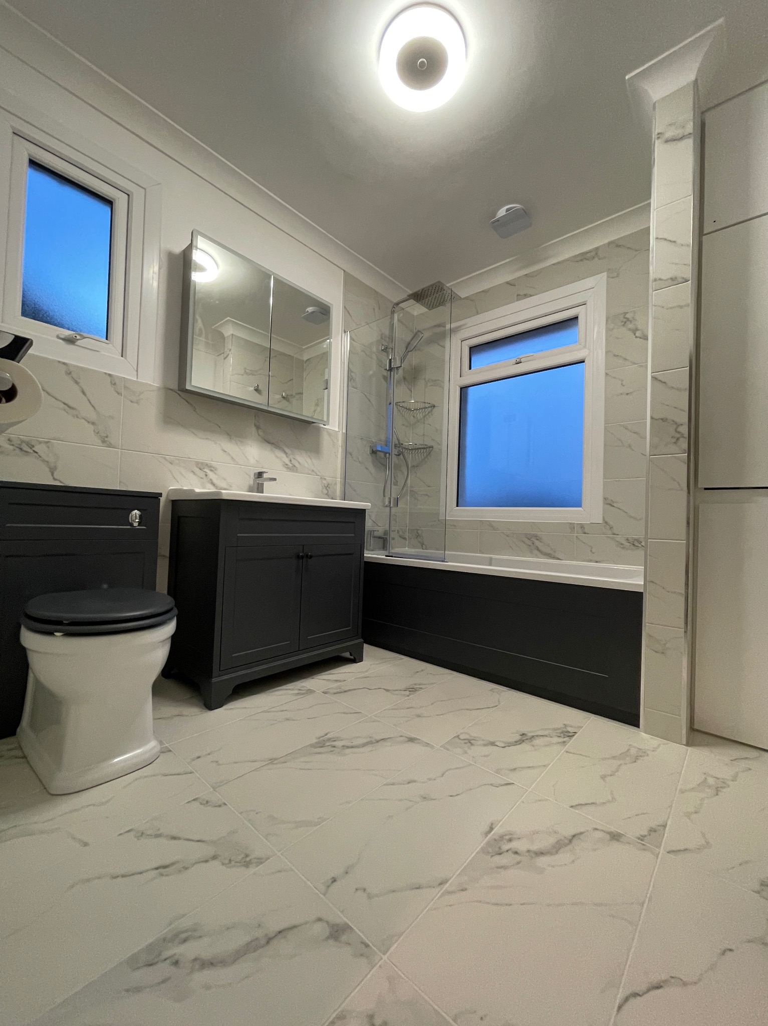 Wykończona łazienka w bieli i szarości z czarnymi akcentami: umywalka z szafką, toaleta i wanna za szklaną ścianką. Marmurowe płytki na podłodze i ścianach.