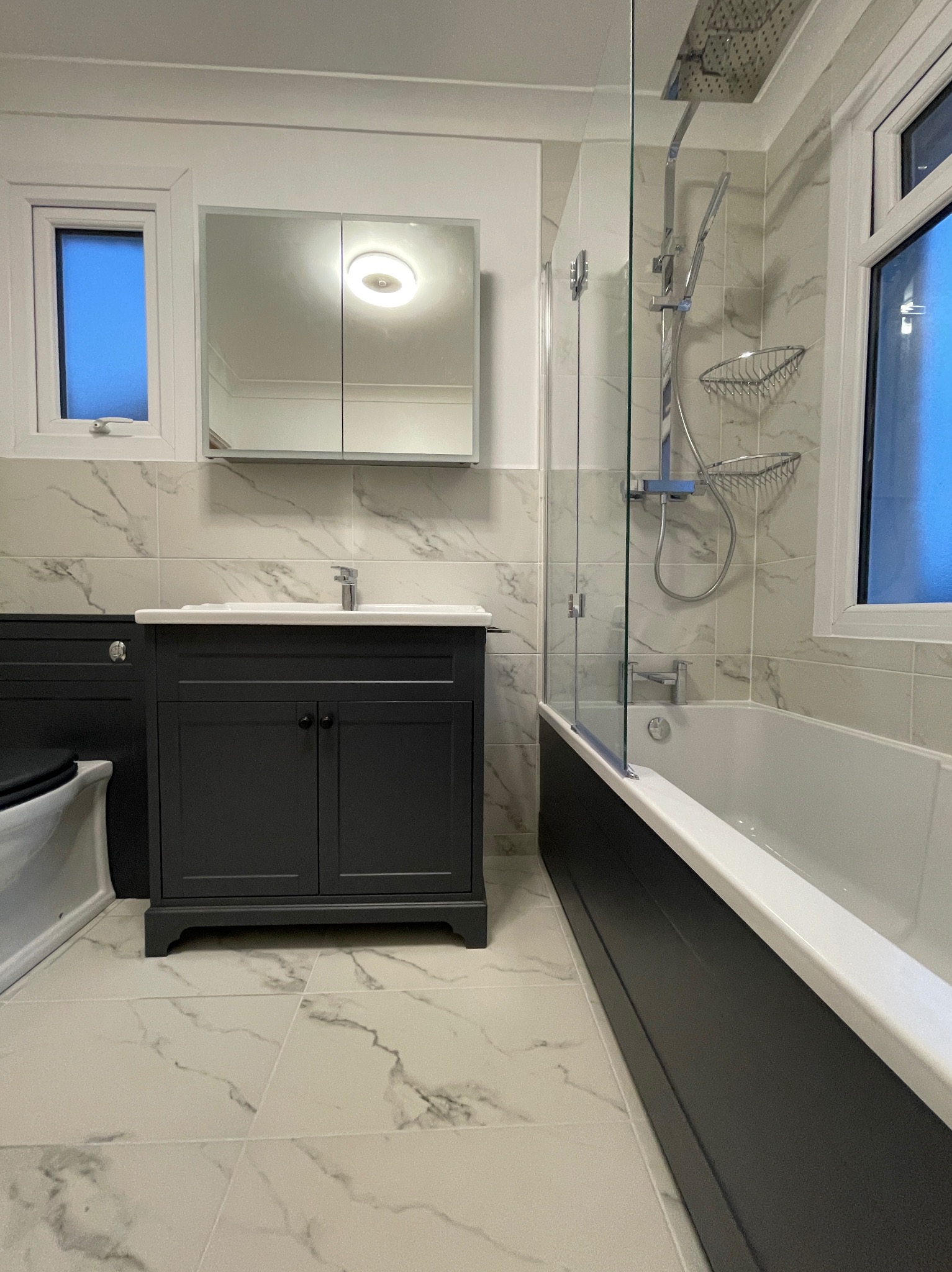 Nowoczesna łazienka z marmurowymi płytkami, grafitową szafką pod umywalką i kabiną prysznicową ze szklaną ścianką. Widoczne okno z niebieskim światłem.