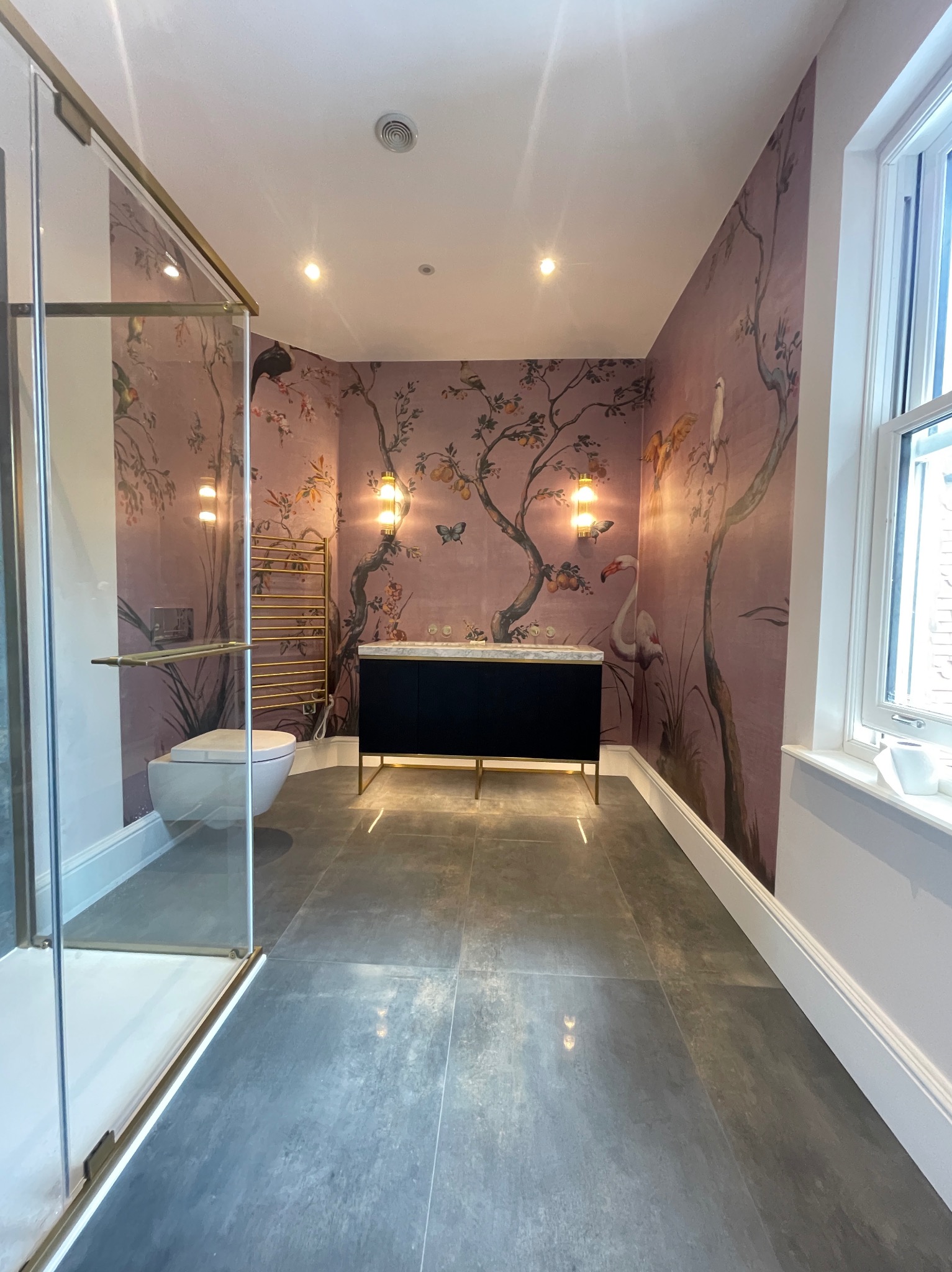 Stylowa łazienka z kabiną prysznicową ze złotymi detalami, toaletą wiszącą i umywalką na tle ściany z motywem drzew i ptaków. Szare płytki podłogowe.