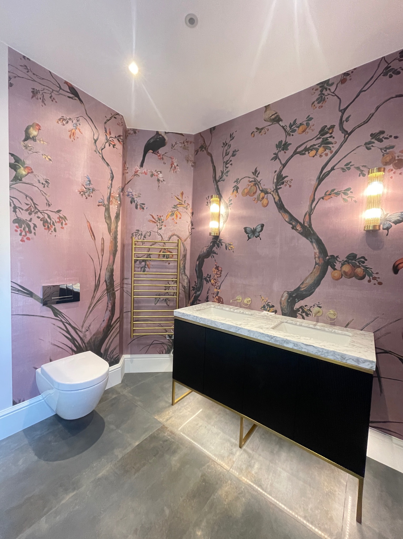 Stylowa łazienka z tapetą w motywy drzew i ptaków. Czarna szafka z marmurowym blatem, złoty grzejnik i szara podłoga tworzą elegancki wystrój.