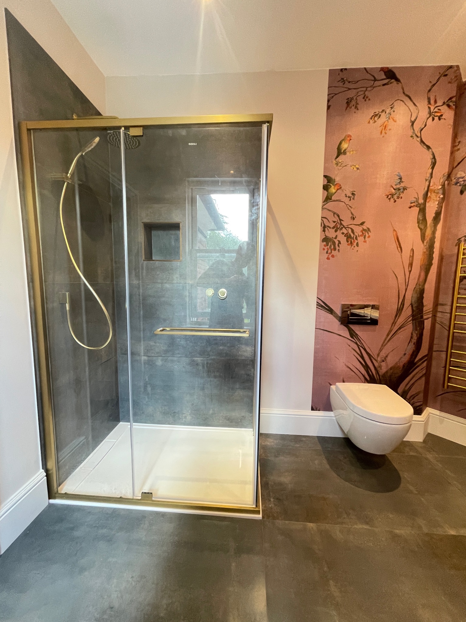 Stylowa łazienka z kabiną prysznicową w złotej ramie, grafitowymi płytkami, toaletą i tapetą w kwiaty i ptaki. Nowoczesny design z dbałością o detale.