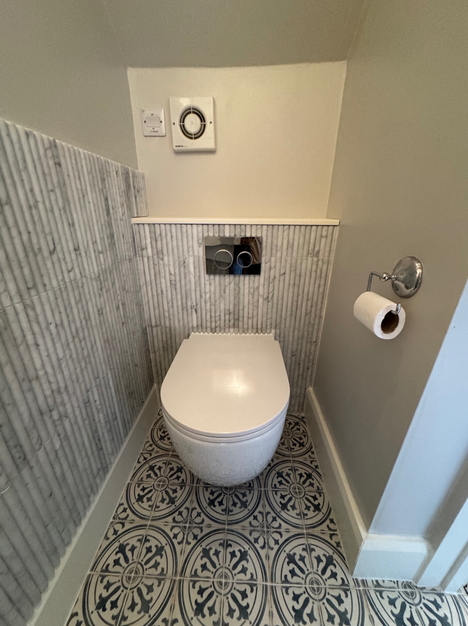 Nowoczesna toaleta podwieszana w łazience wykończonej marmurowymi płytkami i dekoracyjną podłogą. Widoczny wentylator i uchwyt na papier toaletowy.