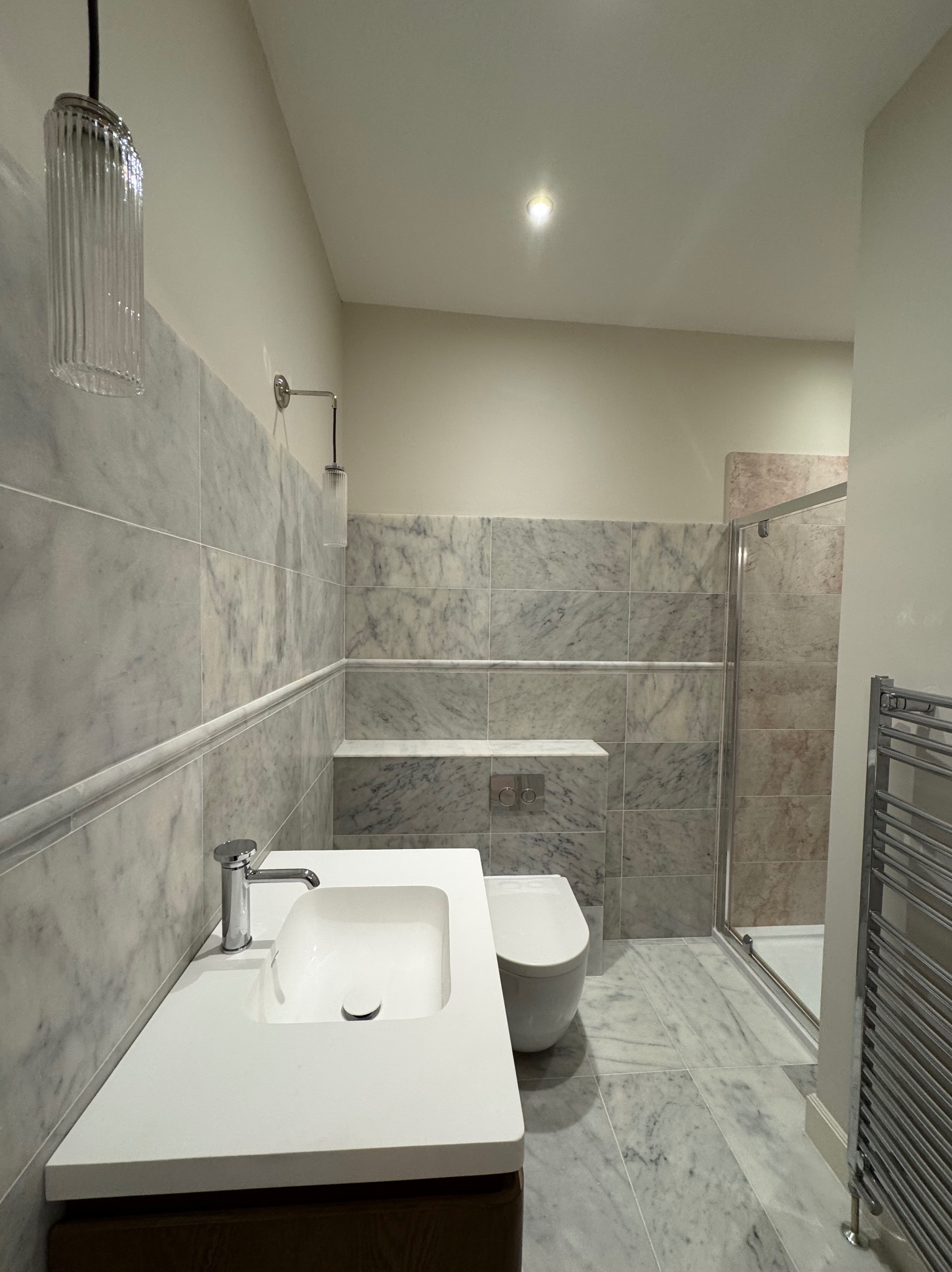 Elegancka łazienka z marmurowymi płytkami, nowoczesną umywalką i szklaną kabiną prysznicową. Widoczne oświetlenie dekoracyjne i grzejnik drabinkowy.