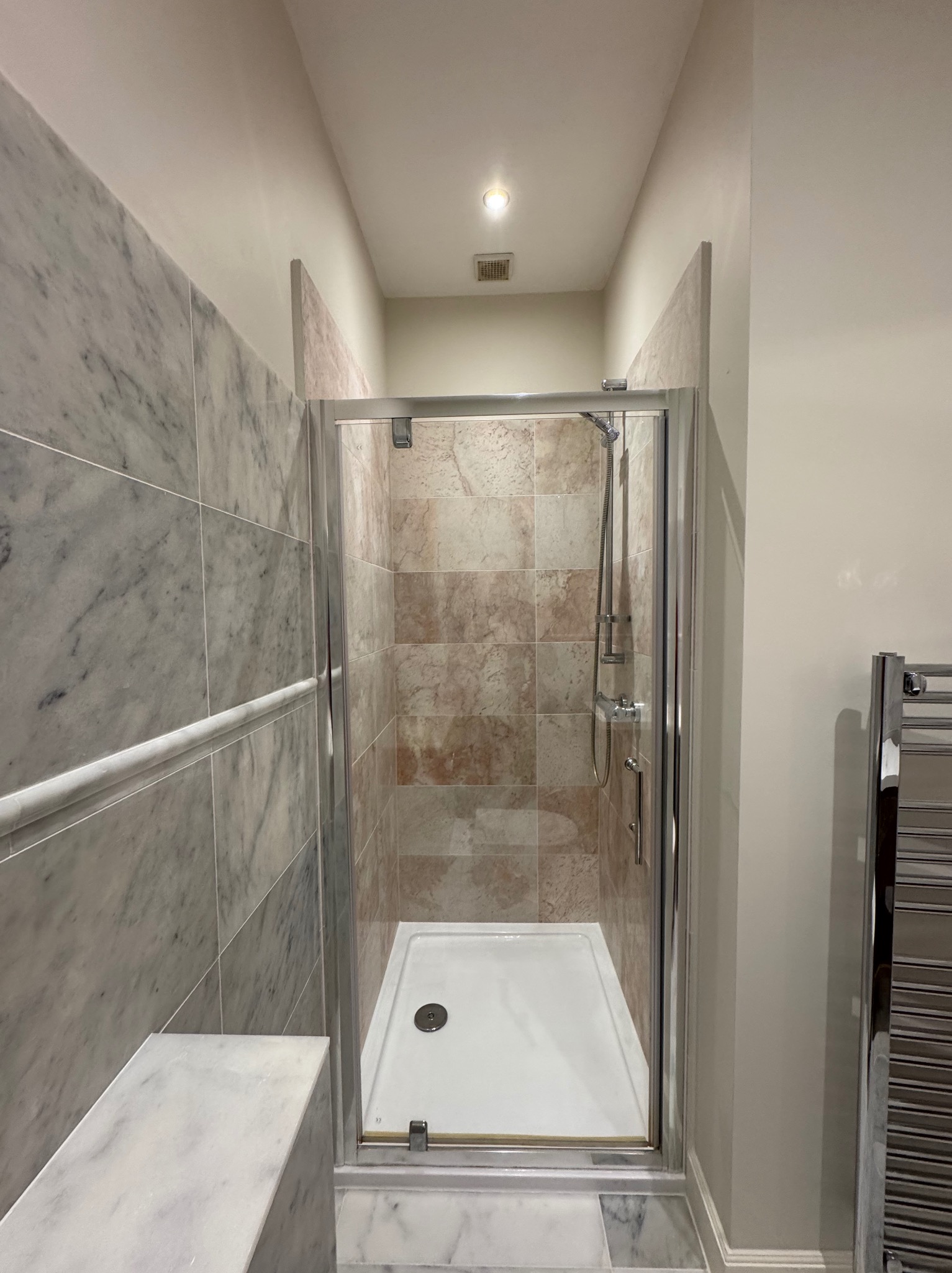 Kabina prysznicowa z marmurowymi płytkami w odcieniach szarości i beżu. Widoczny chromowany panel grzejnikowy i oświetlenie sufitowe.