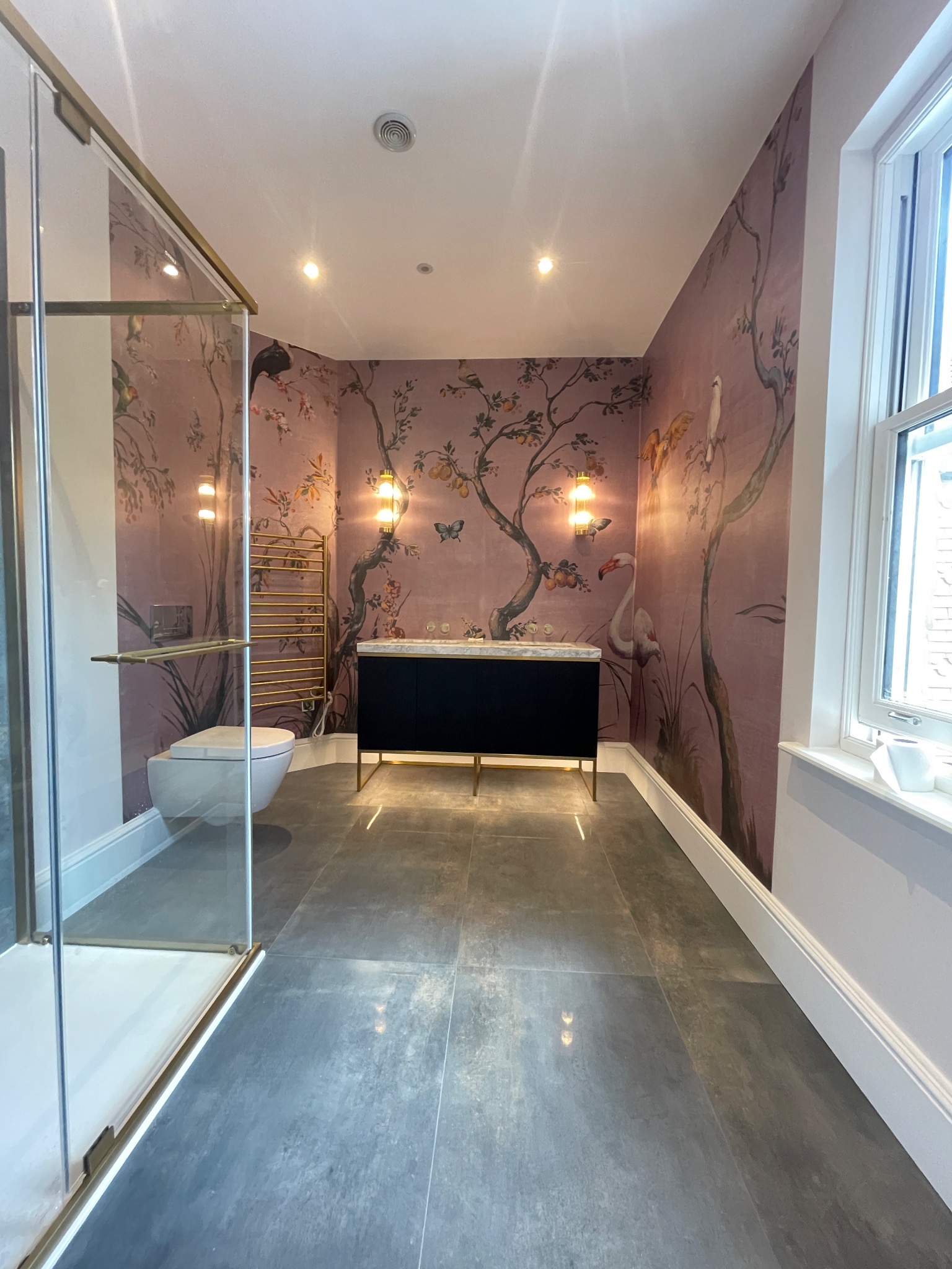 Wykończona łazienka z szaro-grafitową podłogą, kabiną prysznicową ze złotymi detalami i tapetą w motywy roślinne i zwierzęce. Centralnie umieszczona szafka z umywalką.