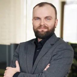 Portret brodatego mężczyzny w szarym garniturze i czarnej koszuli, z założonymi rękami, stojącego w nowoczesnym biurze z przeszkloną ścianą i jasnym oświetleniem.