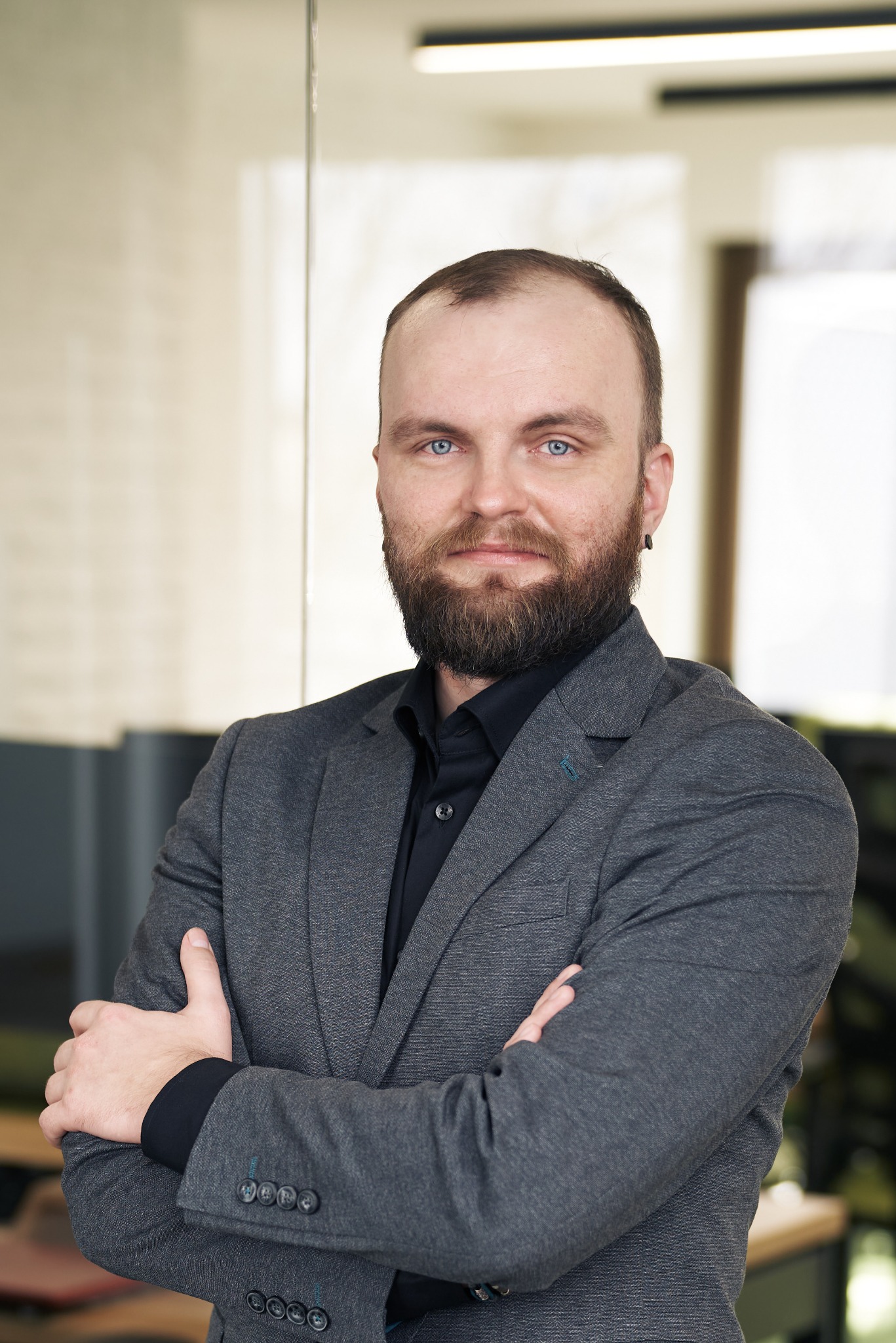 Portret brodatego mężczyzny w szarym garniturze i czarnej koszuli, z założonymi rękami, stojącego w nowoczesnym biurze z przeszkloną ścianą i jasnym oświetleniem.