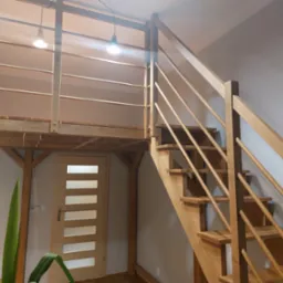 Drewniane schody z balustradą prowadzą na antresolę z drewnianą podłogą. Widoczna lampa sufitowa i drzwi z przeszkleniami.