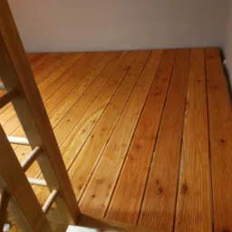 Jasnobrązowa antresola z drewnianą drabinką w jasnym wnętrzu. Deski ułożone równolegle, widoczne sęki i faktura drewna. Ujęcie z dołu, podkreślające solidność konstrukcji.