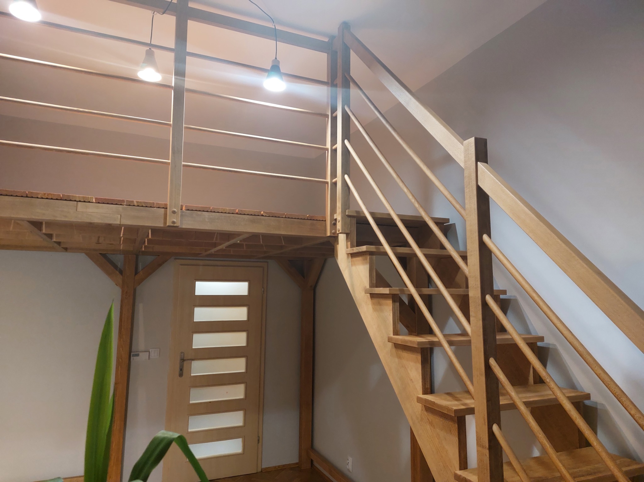 Drewniane schody z balustradą prowadzą na antresolę z drewnianą podłogą. Widoczna lampa sufitowa i drzwi z przeszkleniami.