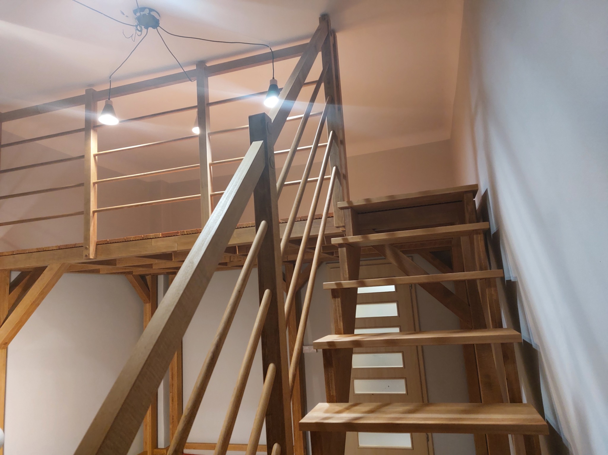 Drewniana antresola z schodami i balustradą, oświetlona lampkami. Widoczne elementy konstrukcyjne i wykończeniowe, jasne drewno. Ujęcie od dołu, podkreślające wysokość konstrukcji.