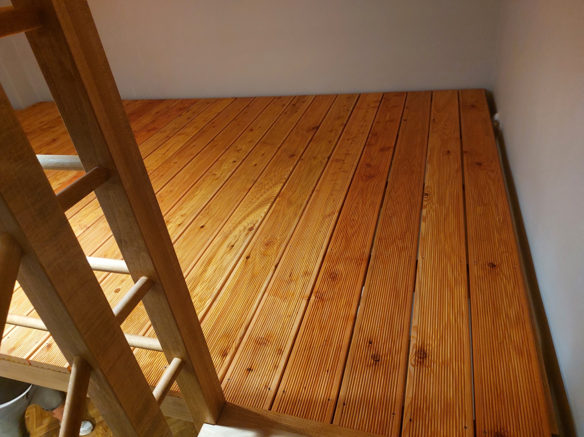 Jasnobrązowa antresola z drewnianą drabinką w jasnym wnętrzu. Deski ułożone równolegle, widoczne sęki i faktura drewna. Ujęcie z dołu, podkreślające solidność konstrukcji.