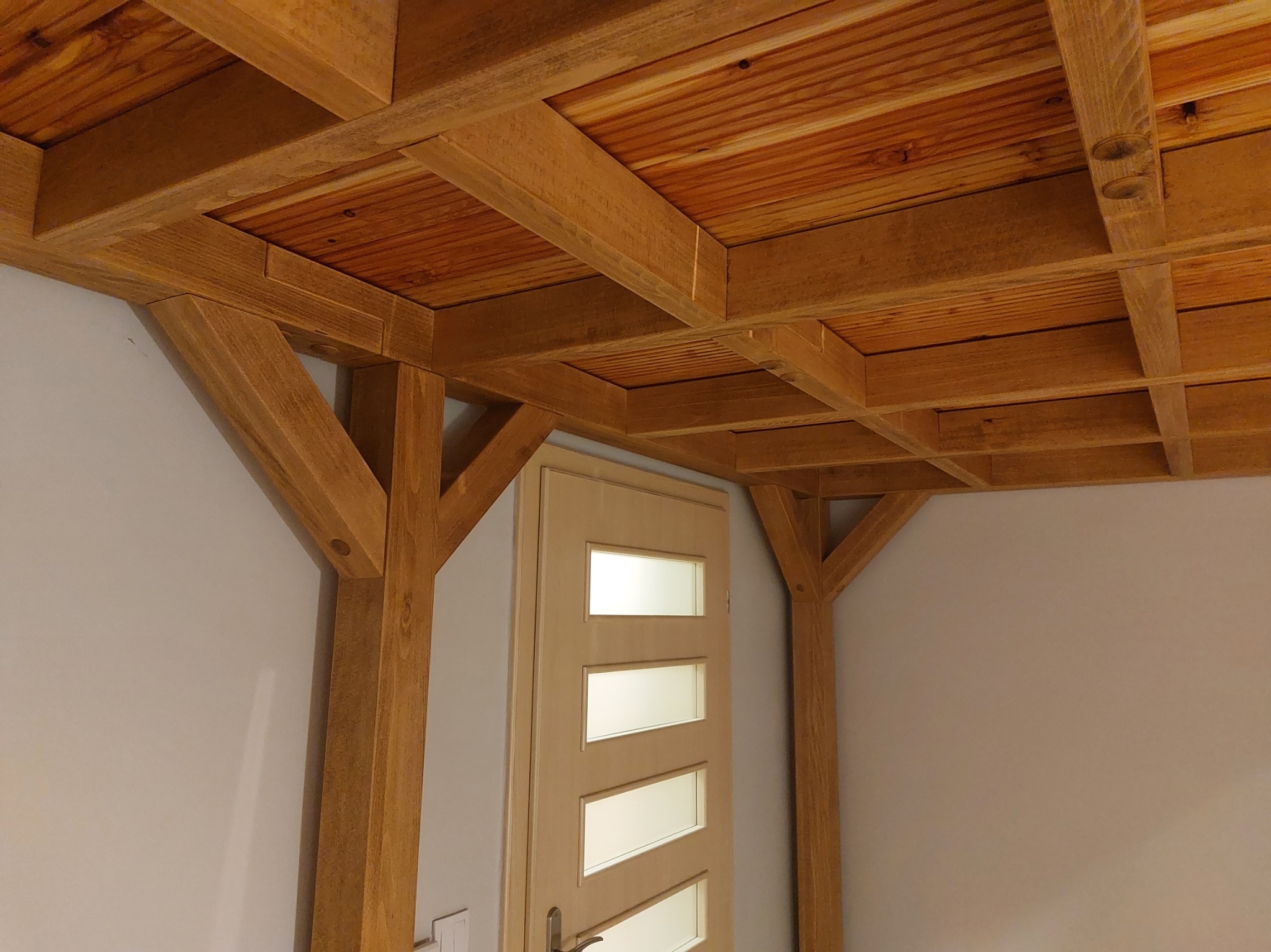 Drewniana konstrukcja antresoli z belek i słupów, widok od dołu. Naturalne drewno, jasne ściany, widoczny fragment drzwi. Solidne wykonanie.