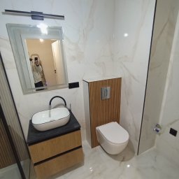 VOLODYMYR ZHURAVEL - Nowoczesna łazienka z marmurowymi płytkami, umywalką nablatową na szafce z drewnianymi lamelami i podwieszaną toaletą z ukrytym systemem spłukiwania, widoczne odbicie osoby robiącej zdjęcie w lustrze.