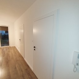 VOLODYMYR ZHURAVEL - Nowoczesne wnętrze z drewnopodobną podłogą, białymi ścianami i dwoma ukrytymi drzwiami z czarnymi klamkami; widoczny wideodomofon na ścianie.