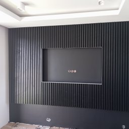 VOLODYMYR ZHURAVEL - Ściana z czarnych pionowych paneli dekoracyjnych z wnęką na telewizor, widoczne wystające kable elektryczne, biała ściana boczna z gniazdkiem elektrycznym w trakcie montażu.