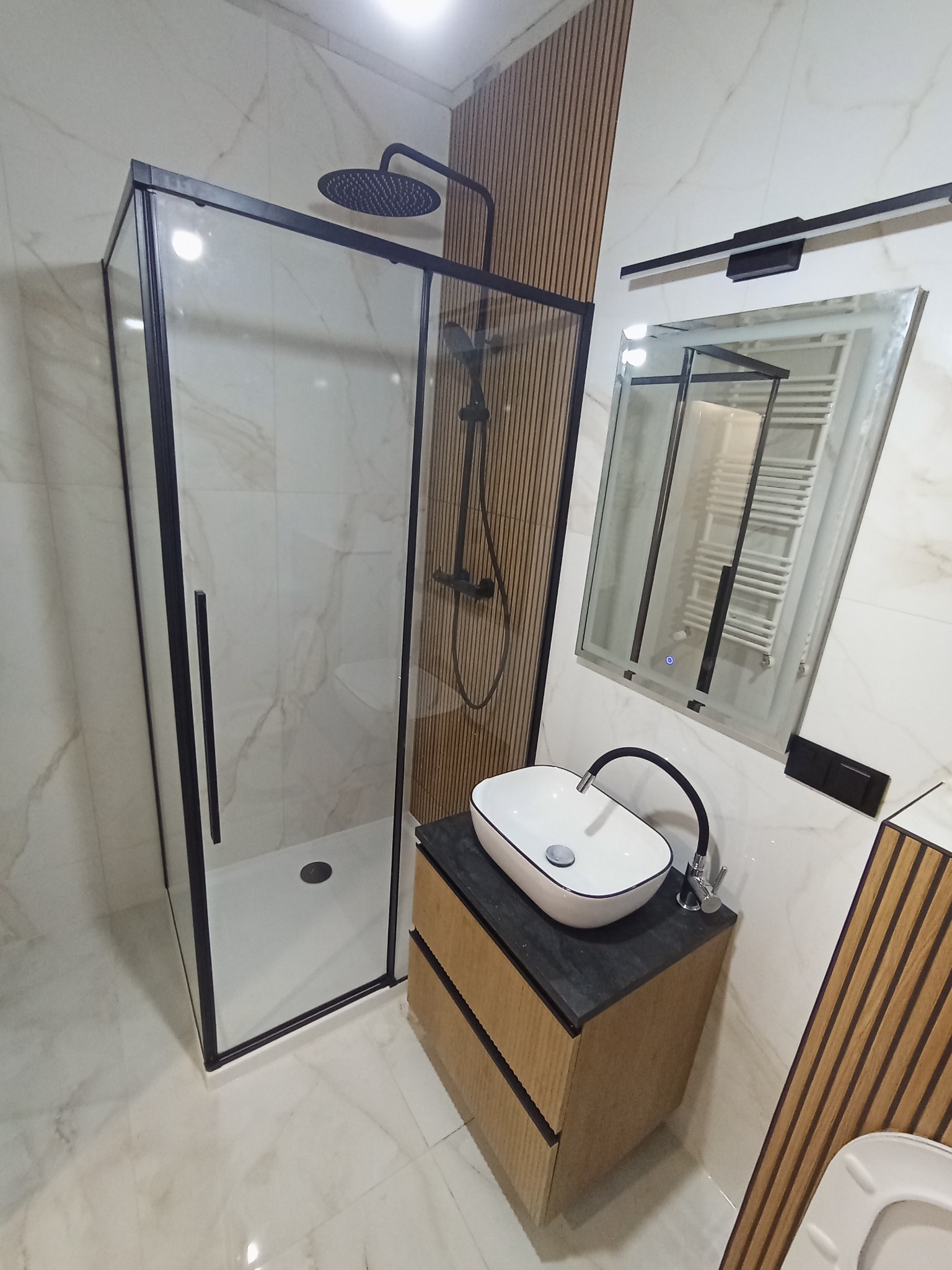 Nowoczesna łazienka z kabiną prysznicową z czarnymi ramami, umywalką nablatową na szafce z drewnianymi lamelami i lustrem z podświetleniem LED.
