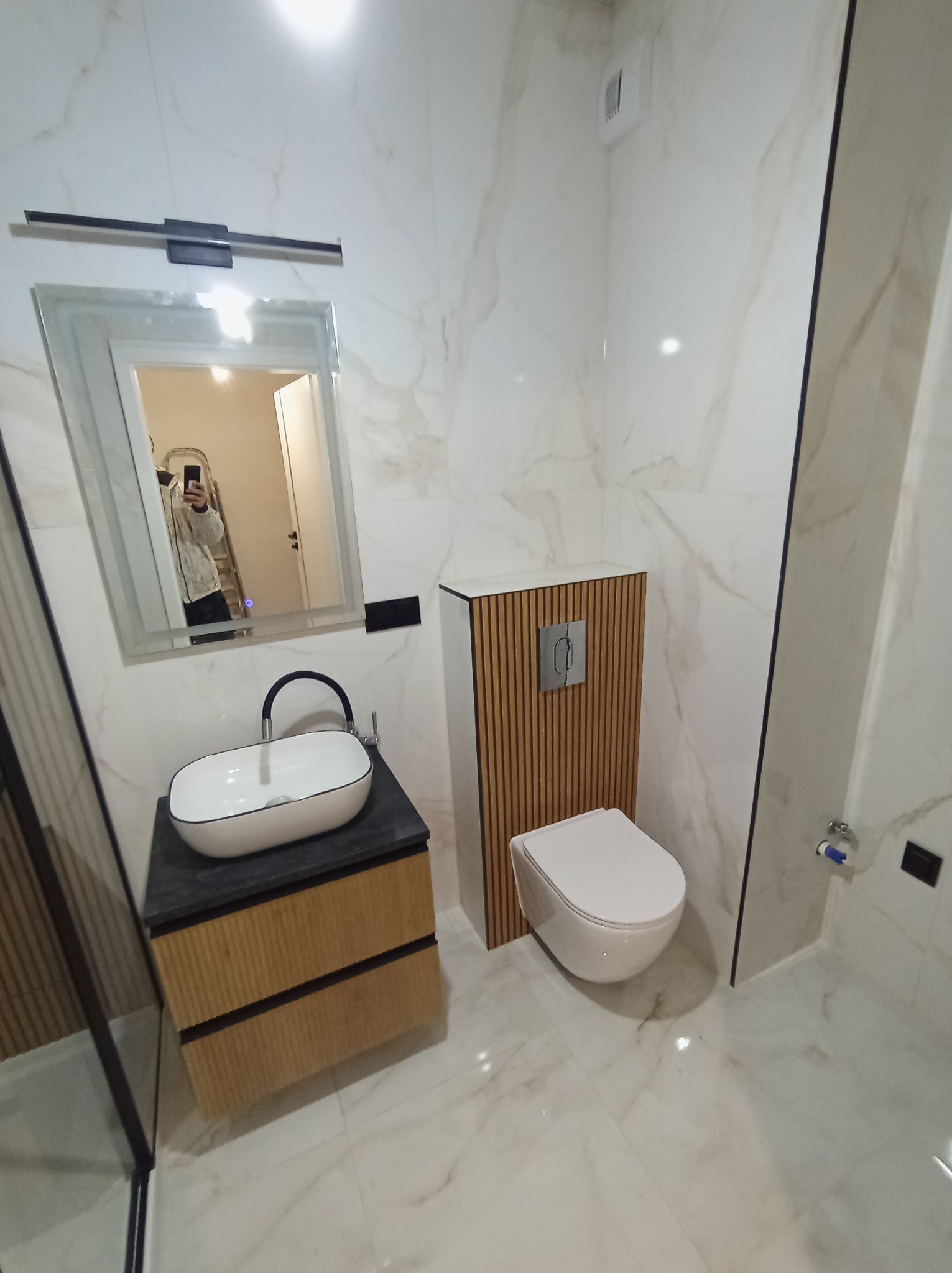 Nowoczesna łazienka z marmurowymi płytkami, umywalką nablatową na szafce z drewnianymi lamelami i podwieszaną toaletą z ukrytym systemem spłukiwania, widoczne odbicie osoby robiącej zdjęcie w lustrze.