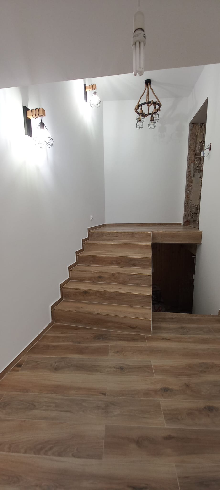 Wykończone schody z drewnopodobnych płytek prowadzące na półpiętro, oświetlone trzema różnymi typami lamp: kinkietami, żyrandolem z liny i żarówką energooszczędną.