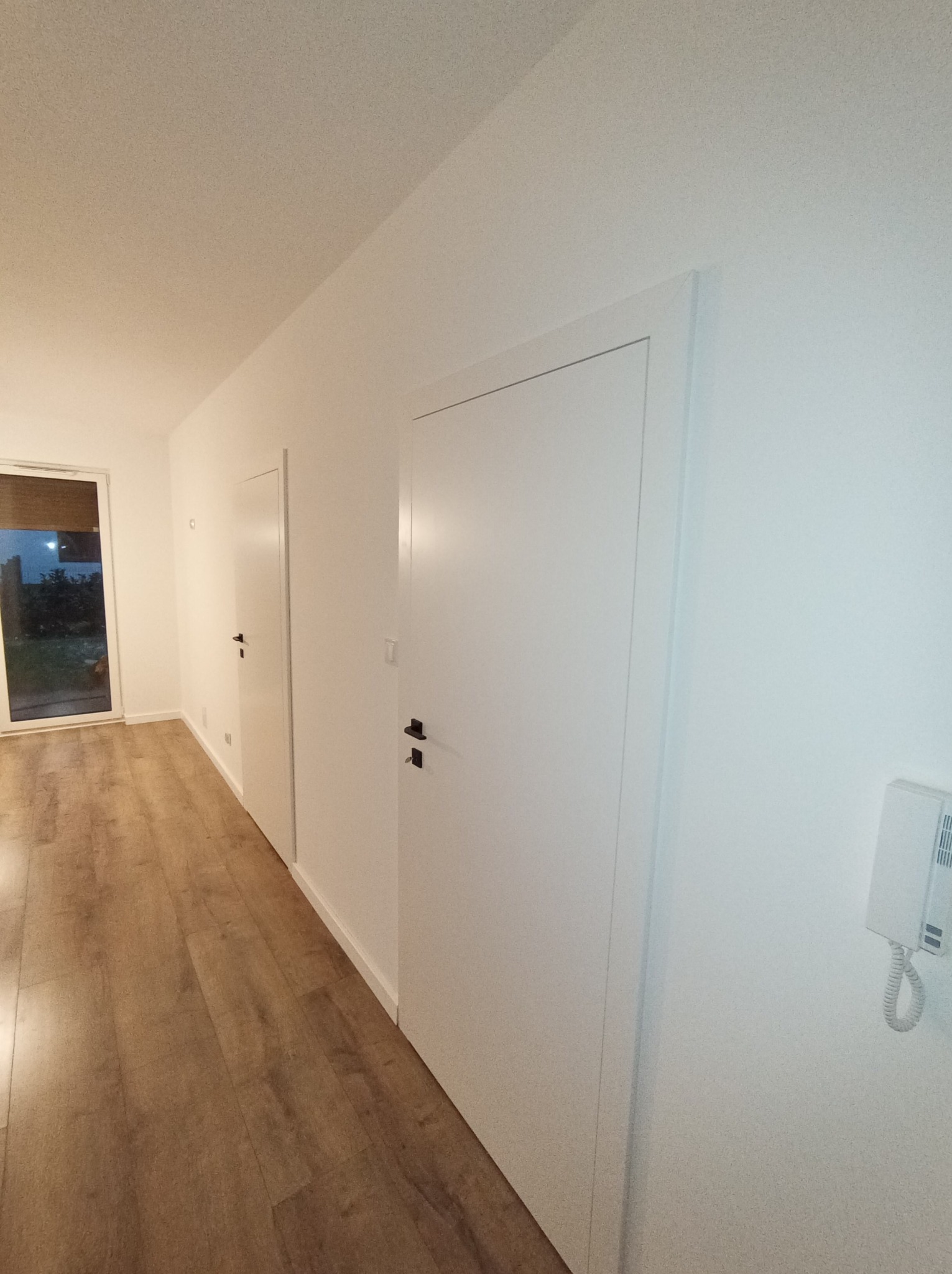 Nowoczesne wnętrze z drewnopodobną podłogą, białymi ścianami i dwoma ukrytymi drzwiami z czarnymi klamkami; widoczny wideodomofon na ścianie.