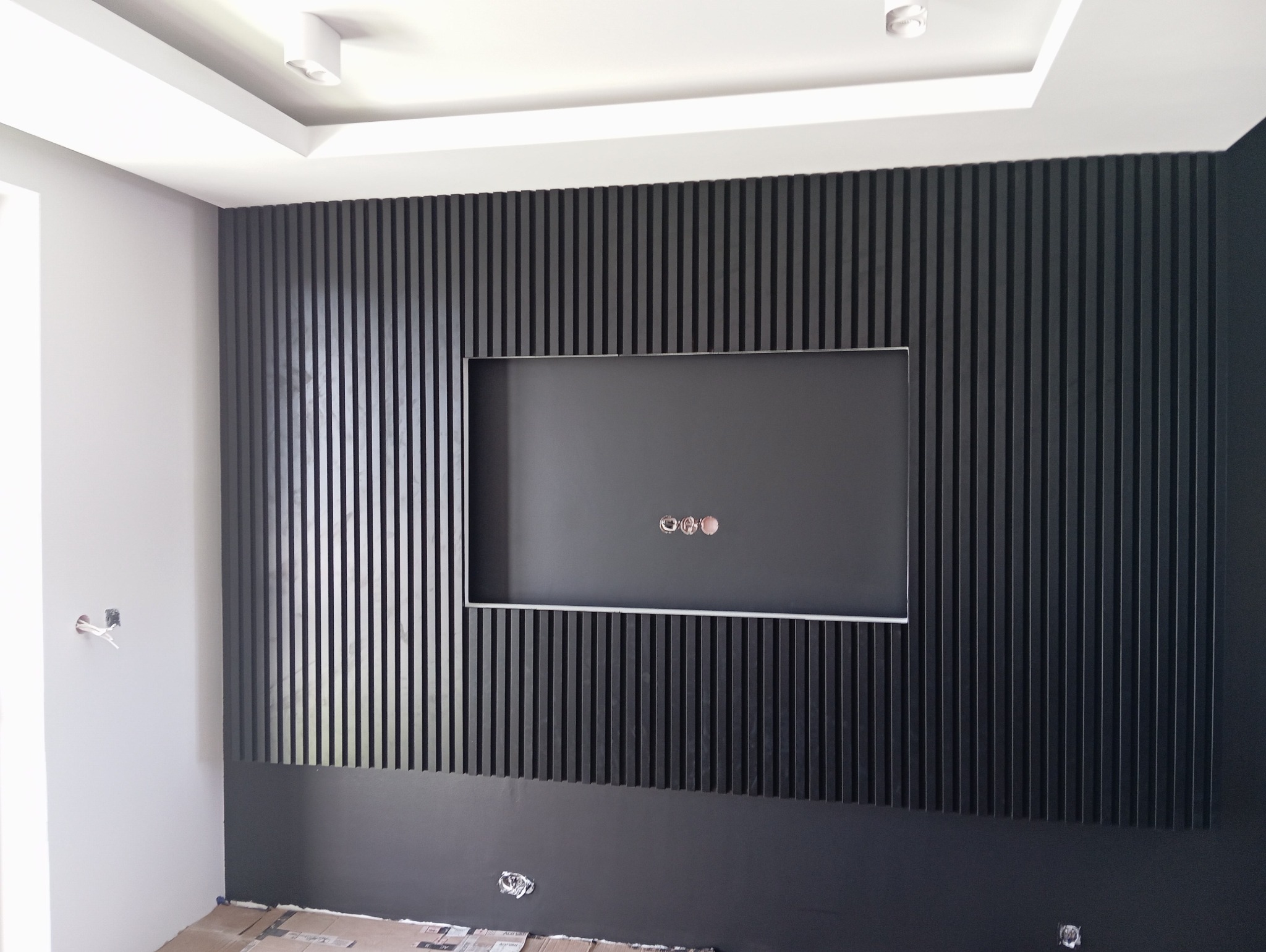 Ściana z czarnych pionowych paneli dekoracyjnych z wnęką na telewizor, widoczne wystające kable elektryczne, biała ściana boczna z gniazdkiem elektrycznym w trakcie montażu.