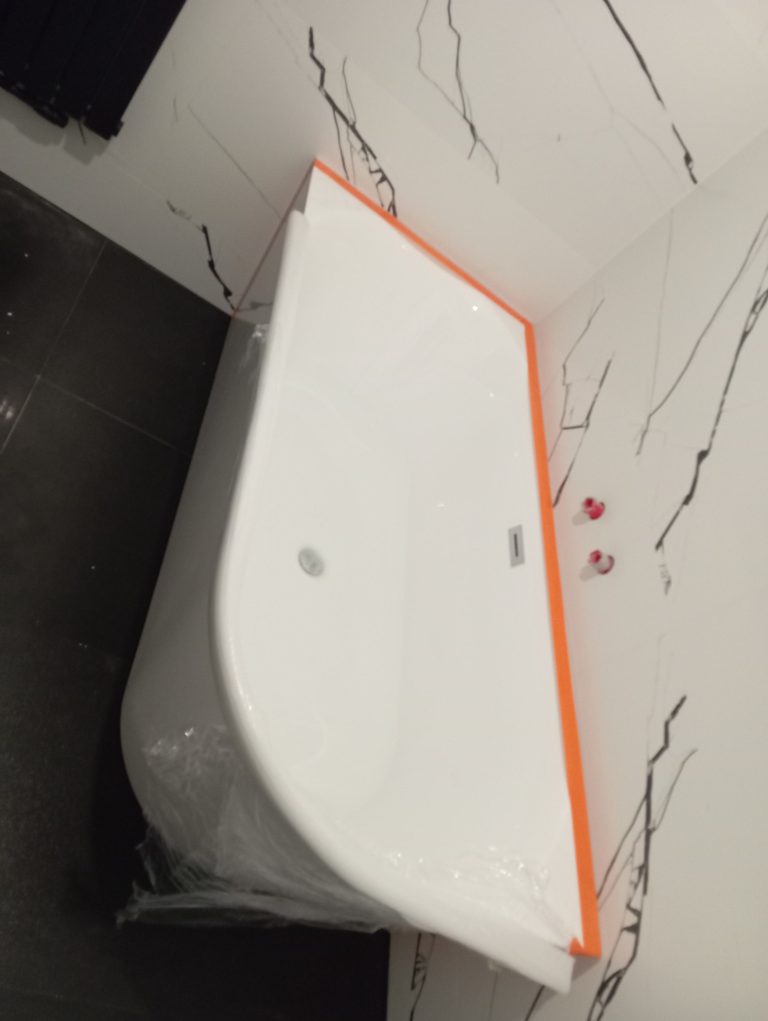 Nowa, biała wanna narożna z pomarańczowym obramowaniem, zabezpieczona folią ochronną, stoi w łazience z czarno-białymi płytkami imitującymi marmur.